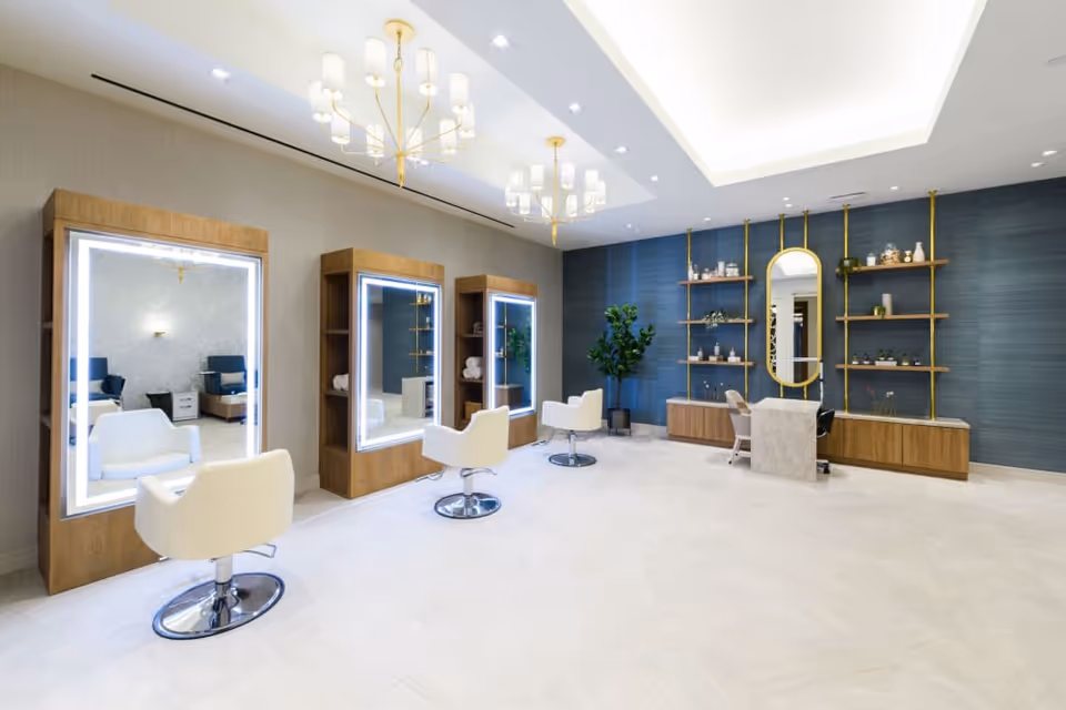 Bright modern salon interior with illuminated styling mirrors, white swivel chairs, shelving and a reception desk.