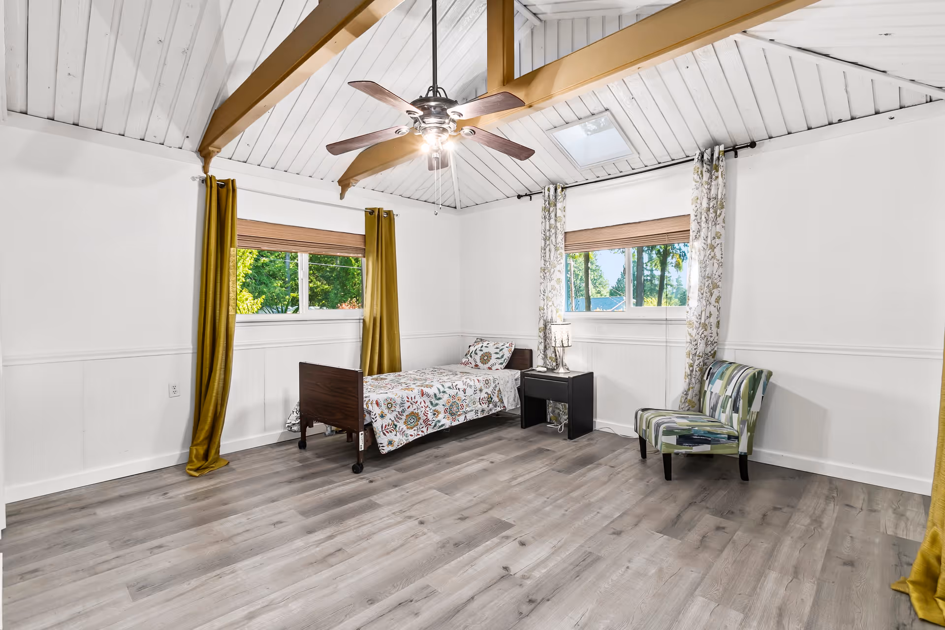 Bright bedroom with a single bed, nightstand, patterned armchair, ceiling fan, skylight and windows with curtains.
