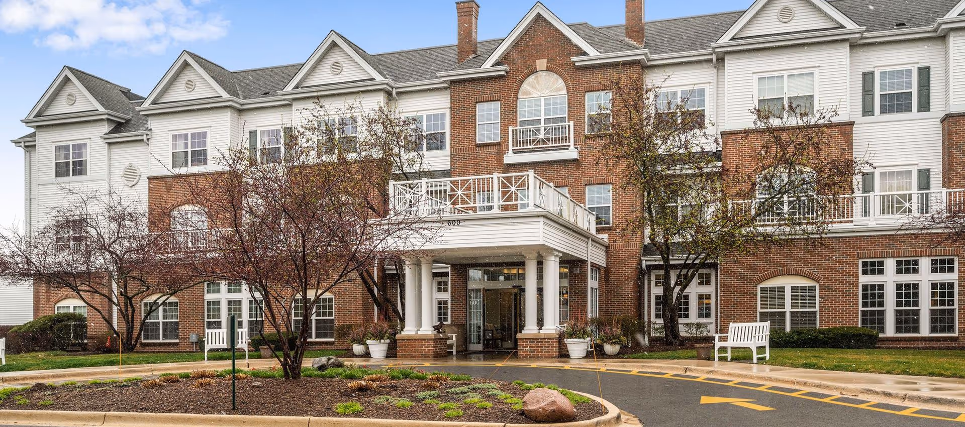 Exterior front view of a senior living facility building with a brick and white siding facade, multiple windows, a covered entrance with white columns, small trees, and a circular driveway with landscaping.