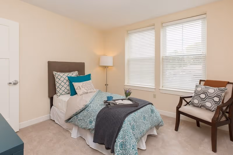 A neatly made single bed with patterned teal and white bedding, multiple pillows, and a dark gray throw blanket in a bright bedroom. A tray with a cup, book, and small vase with flowers is placed on the bed. Next to the bed is a floor lamp and a wooden chair with a patterned cushion. Two large windows with white blinds allow natural light into the room.
