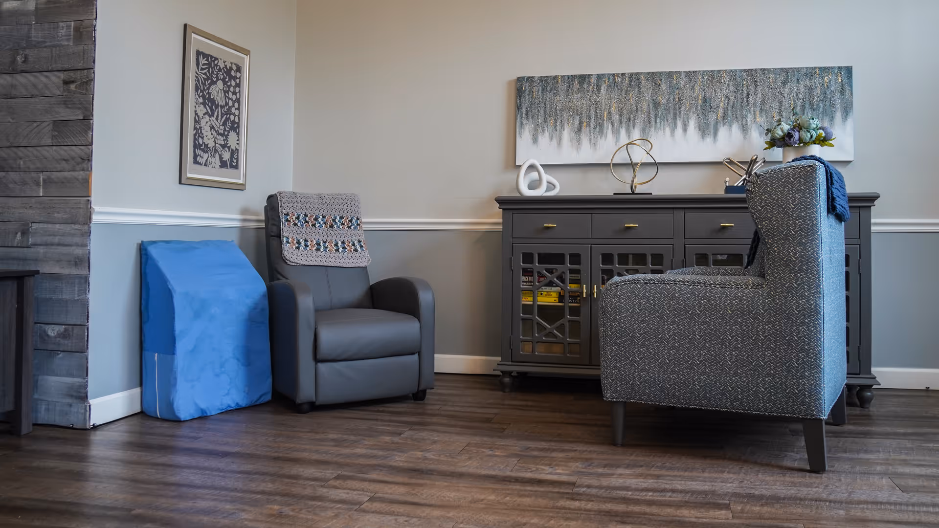 A cozy sitting area with a gray recliner chair draped with a crocheted blanket, a blue wedge cushion, and a patterned armchair facing a dark wooden sideboard. The sideboard has decorative items on top, including abstract sculptures and a floral arrangement. A large abstract painting hangs above the sideboard on a two-tone gray wall with white trim. The floor is wood with a dark finish.