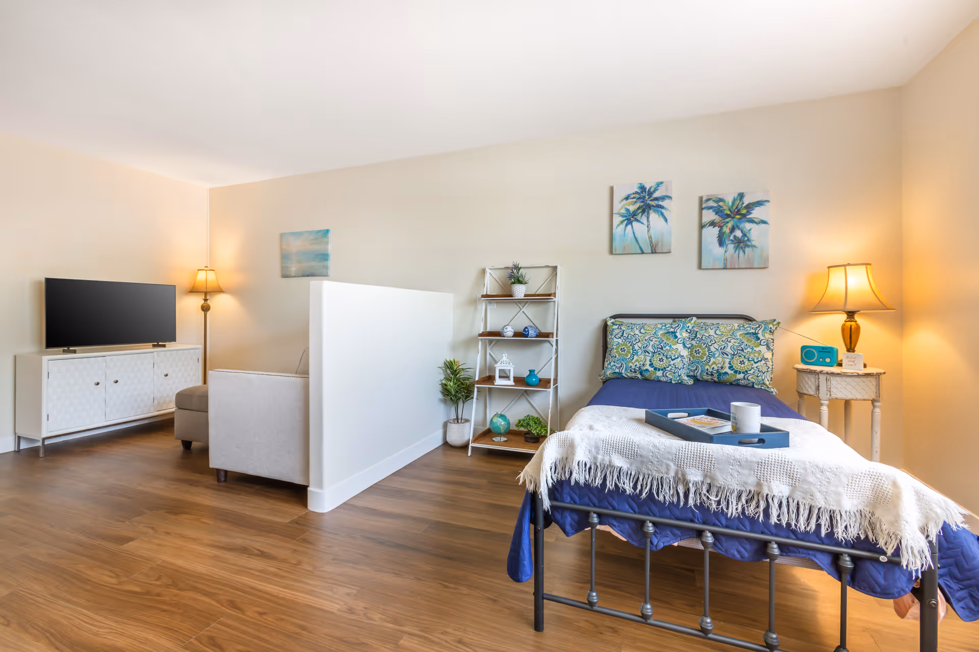 A bright and cozy room featuring a single bed with blue bedding and a white throw blanket, a small side table with a lamp and a blue radio, a shelving unit with decorative items, and a partial view of a living area with a TV on a white cabinet and a floor lamp. The room has wooden flooring and light-colored walls adorned with artwork.