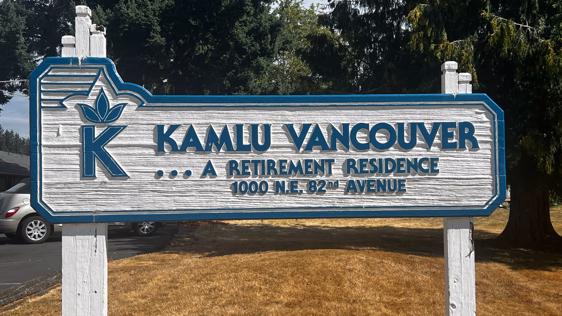 A wooden sign painted white with blue trim and lettering that reads 'KAMLU VANCOUVER ... A RETIREMENT RESIDENCE 1000 N.E. 82nd AVENUE' standing outdoors on a patch of dry grass with trees and parked cars in the background.
