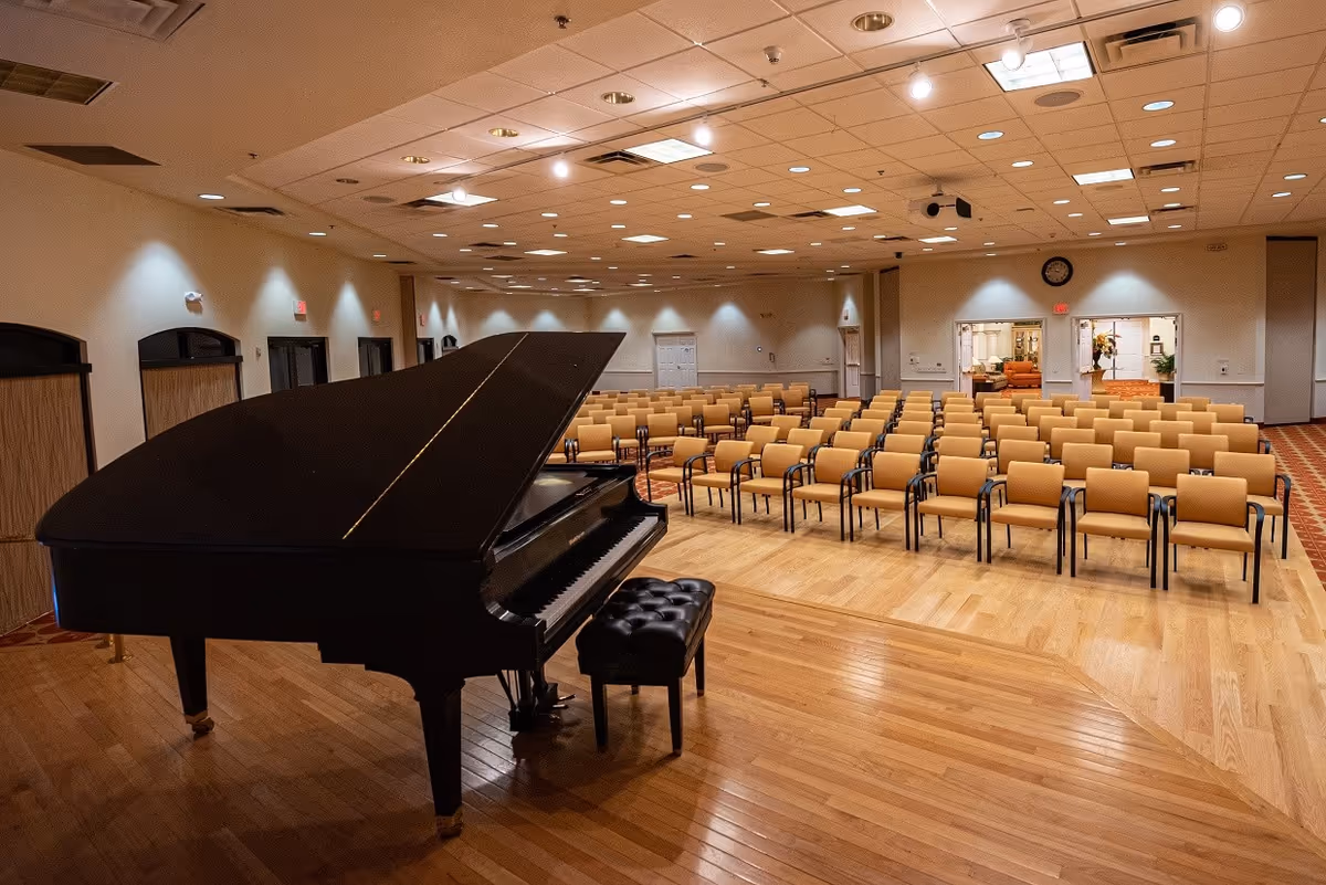 A large room with a wooden floor stage featuring a black grand piano and piano bench. In front of the stage, there are multiple rows of beige chairs arranged for an audience. The room has a high ceiling with many recessed lights and white walls. At the back, there are double doors leading to another room with more seating and decorations.
