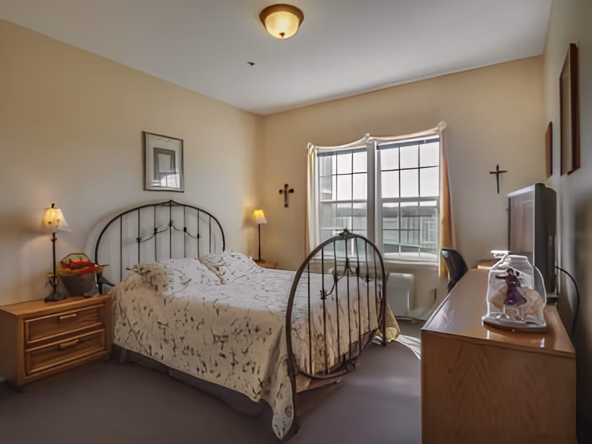 A cozy bedroom featuring a wrought iron bed, bedside lamps, and a wooden dresser. The room has large windows with curtains, allowing natural light to fill the space, and is decorated with framed artwork and a small decorative figure.