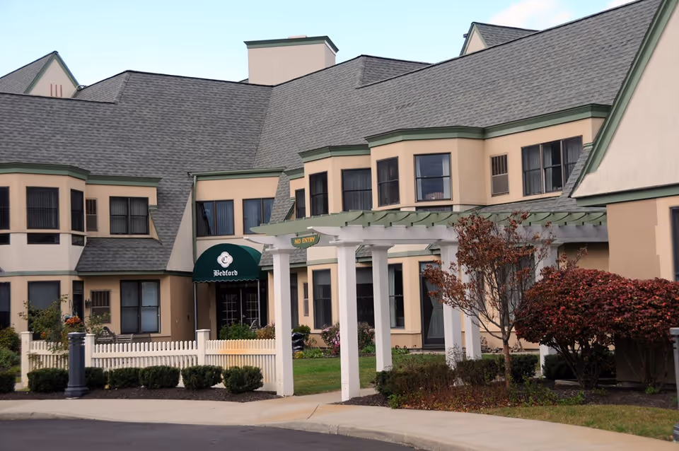 Exterior front of a senior living building with a covered pergola entrance, green awning, beige facade and landscaped shrubs.
