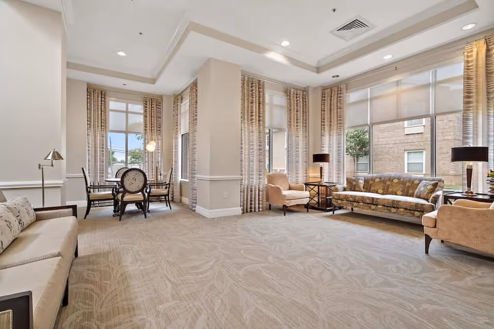 A spacious and well-lit living room area with large windows covered by patterned curtains. The room features a patterned sofa, two beige armchairs, a beige bench, two side tables with lamps, and a small round table with four chairs near the windows. The carpet has a subtle leaf pattern, and the walls are painted in a light neutral color.