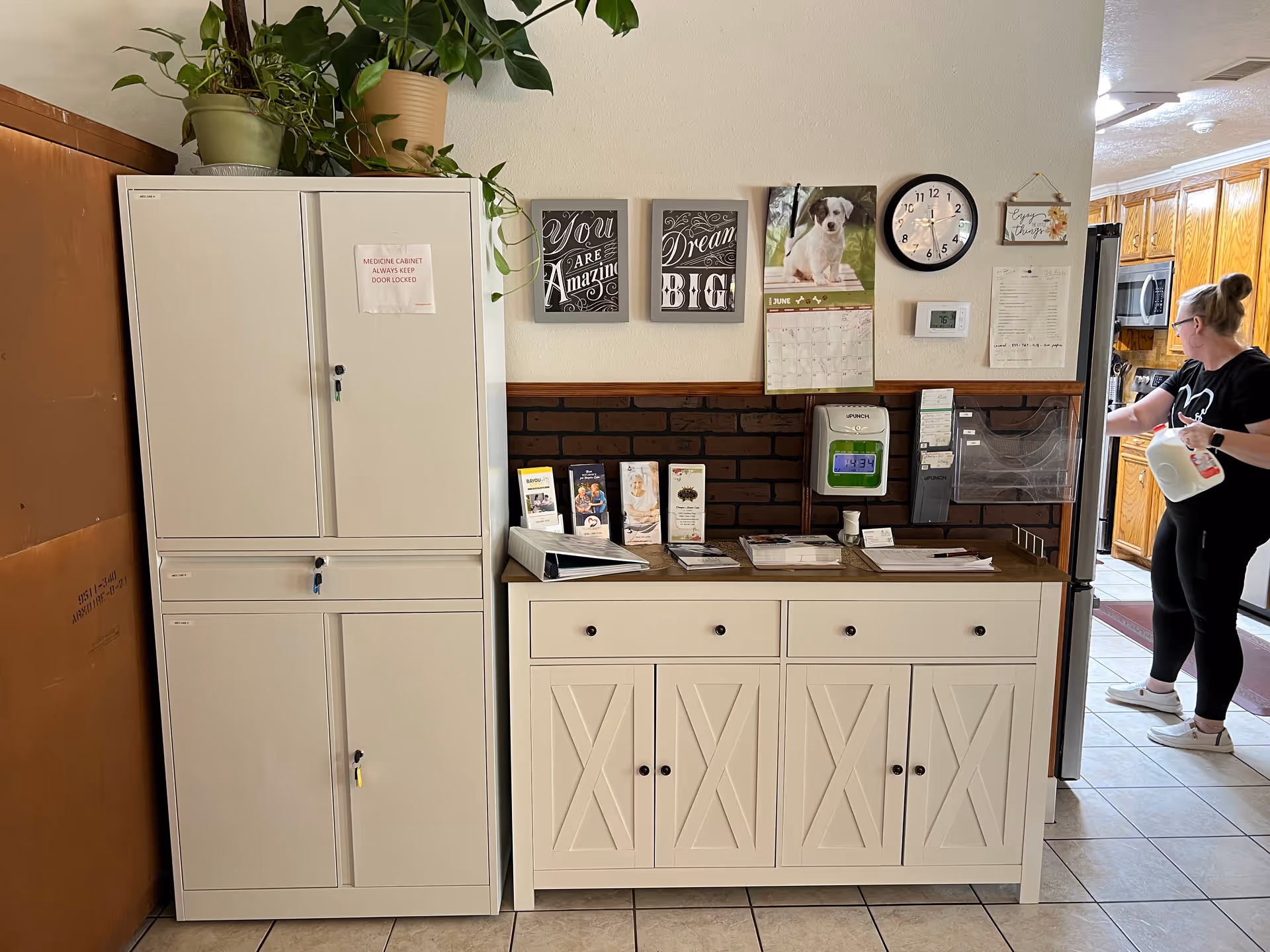 Interior view of a senior care facility area showing a white medicine cabinet with a sign that reads 'MEDICINE CABINET ALWAYS KEEP DOOR LOCKED', a white sideboard with brochures, a time clock device, a calendar with a dog picture, a clock, and motivational wall art. A woman holding a gallon of milk is standing near a kitchen area with wooden cabinets and appliances.