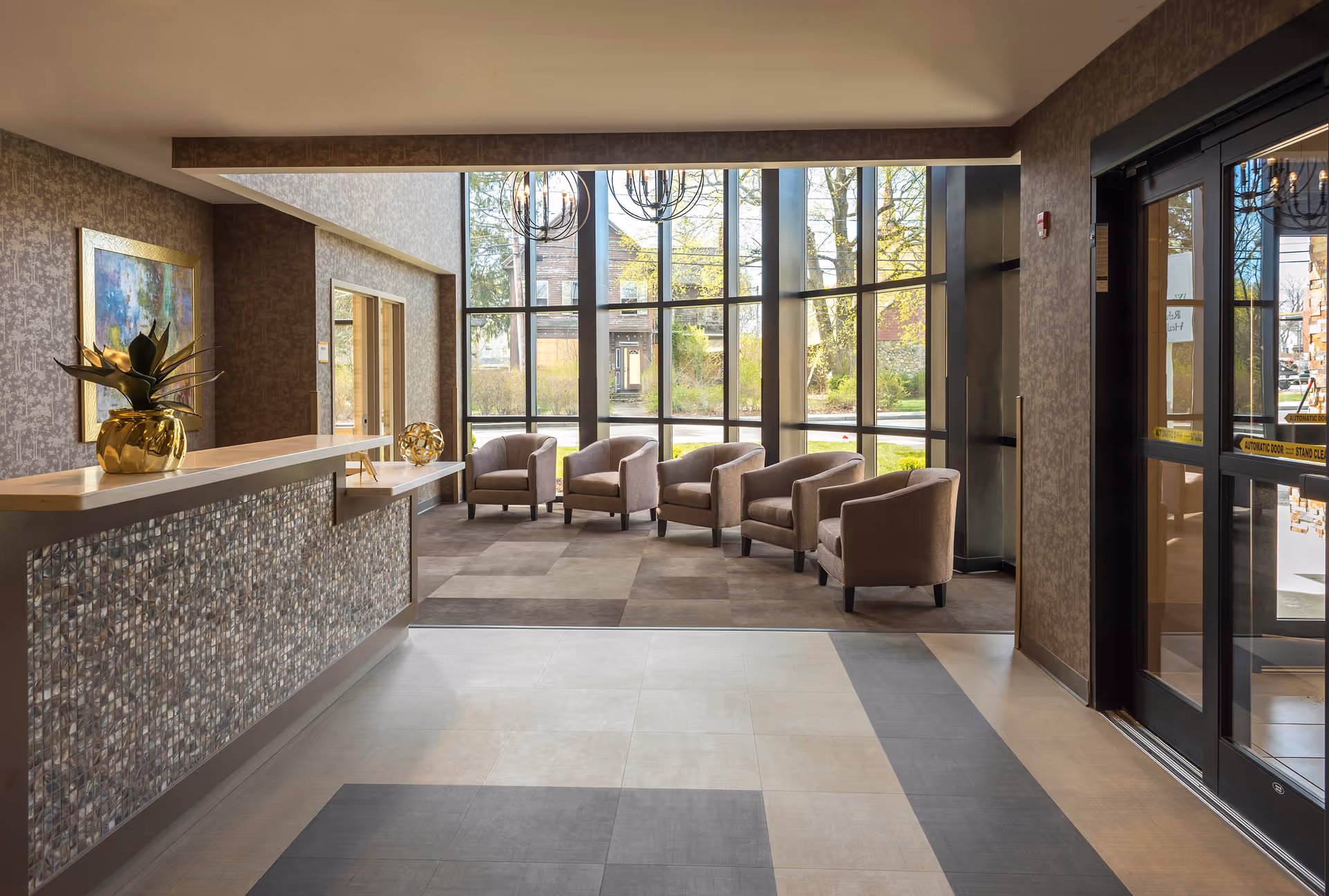 A bright and modern reception area with a decorative front desk on the left, featuring a golden plant pot and a decorative orb. In the background, there is a seating area with six beige armchairs arranged in two rows near large floor-to-ceiling windows that allow natural light to fill the space. The floor has a patterned tile design, and the walls are decorated with textured wallpaper and a framed painting. The entrance door is visible on the right side.
