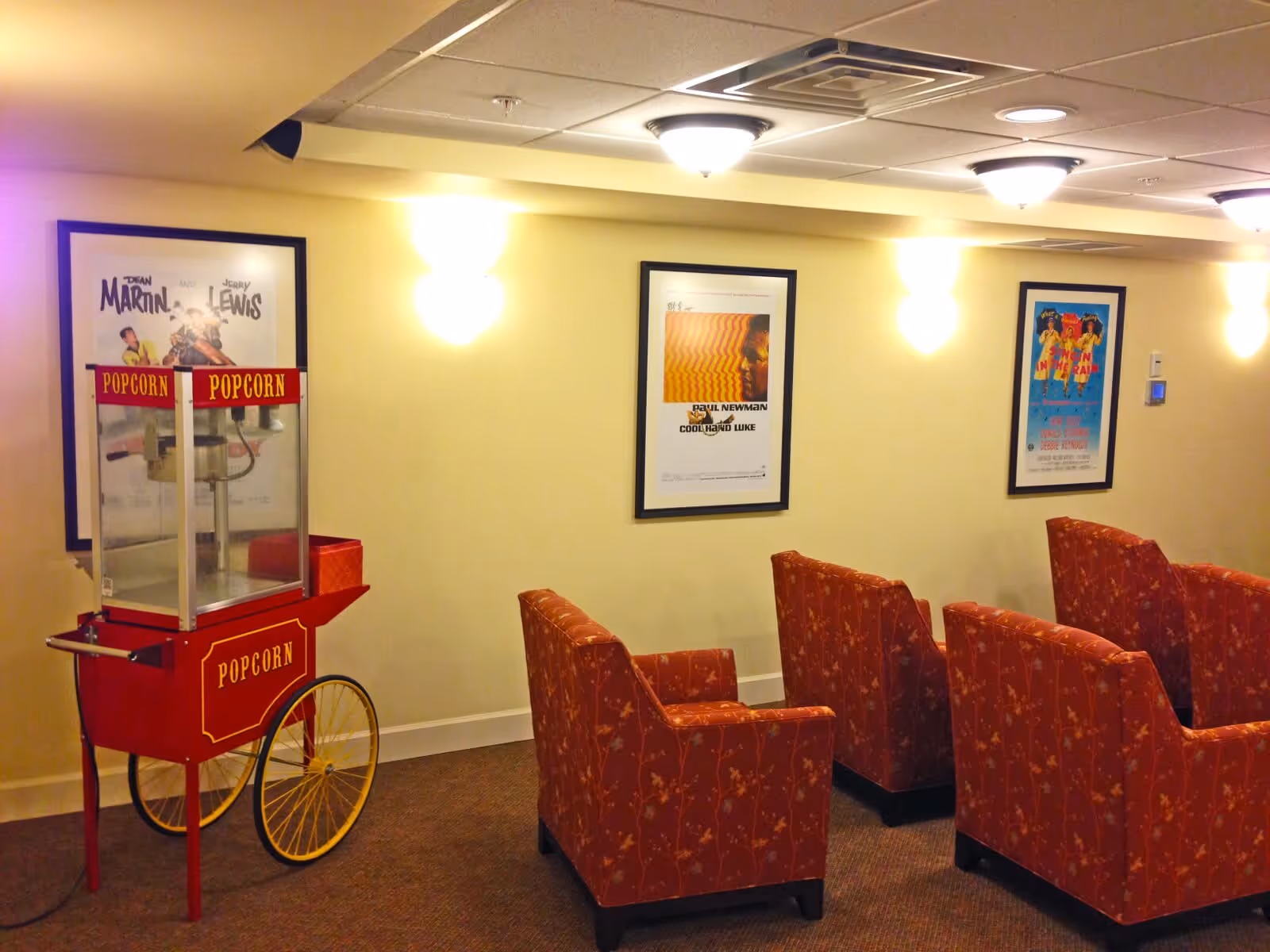 A small theater room with red patterned armchairs arranged in rows facing a screen. On the left side, there is a vintage-style red popcorn machine with yellow wheels. The walls are light yellow and decorated with framed movie posters. The ceiling has recessed lighting fixtures.