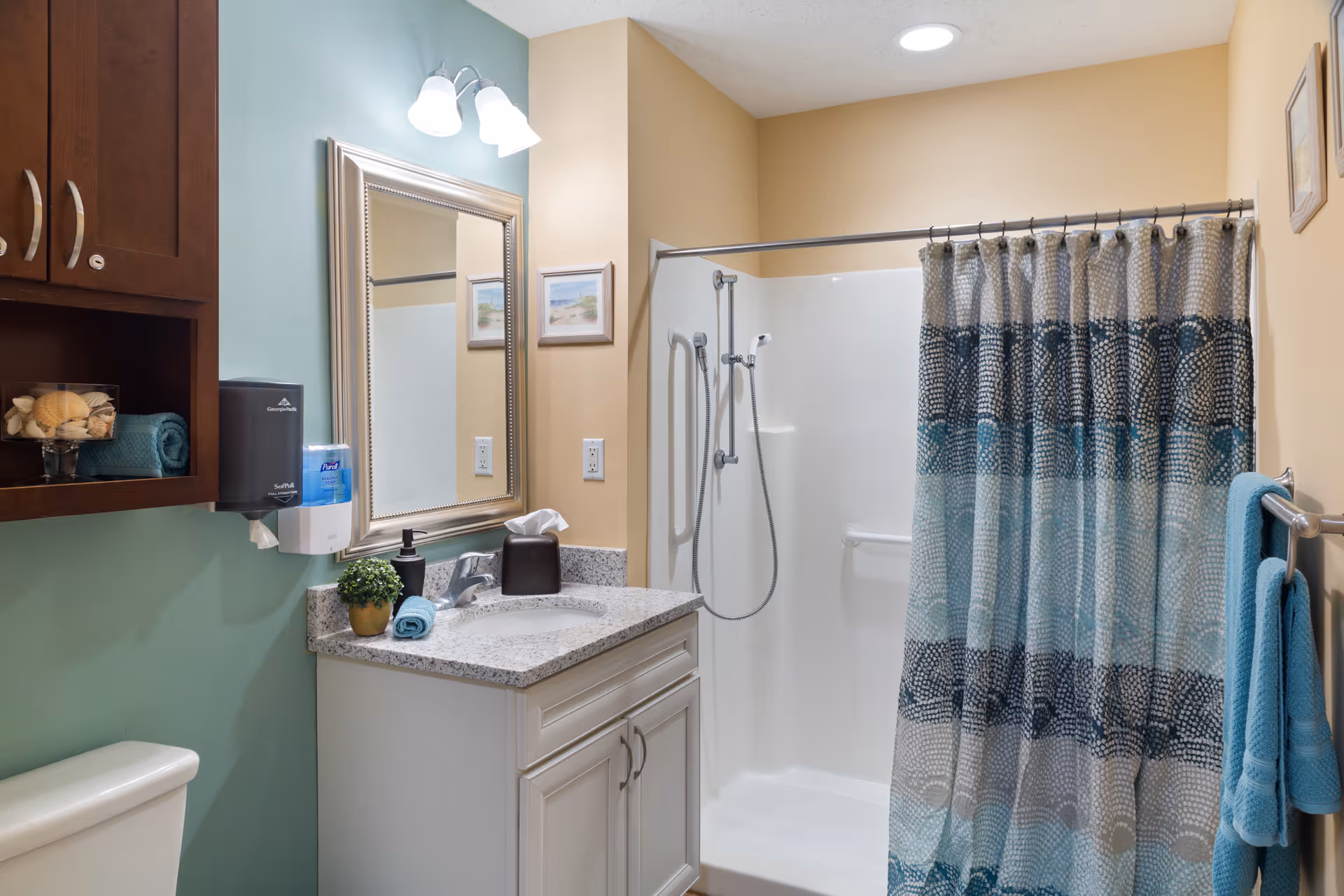 A clean and well-lit bathroom featuring a white vanity with a granite countertop, a mirror with a decorative frame, a shower with a blue and gray patterned curtain, and a toilet. The walls are painted in soft green and beige tones, and there are towels and decorative items neatly arranged.