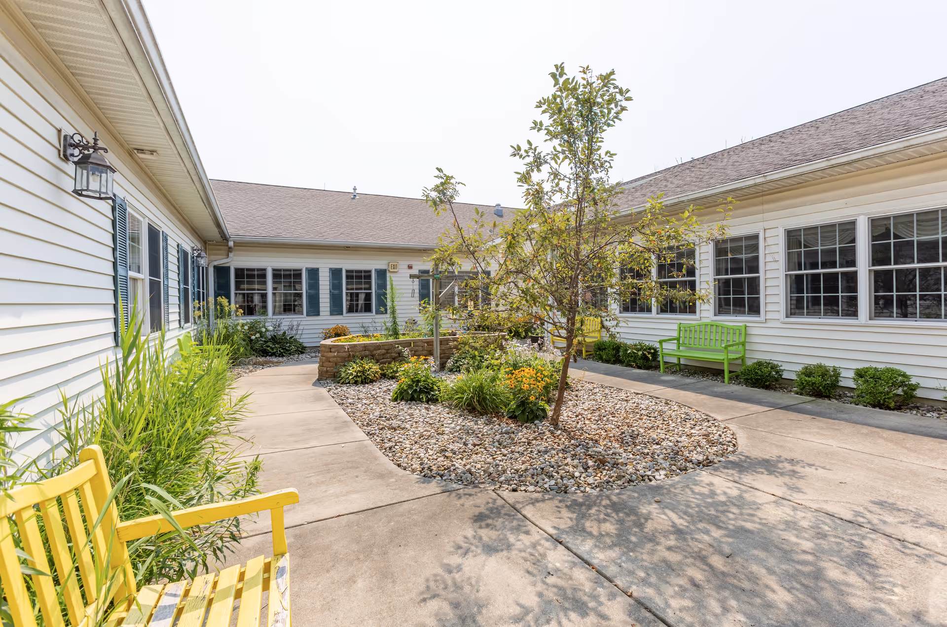 Outdoor courtyard area at StoryPoint Granger with a paved walkway surrounding a landscaped garden bed featuring a small tree and various plants. Yellow and green benches are placed along the walkway next to the white building with multiple windows and blue shutters.