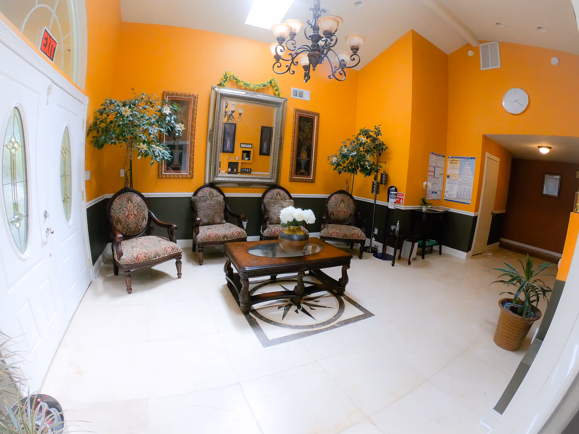 A cozy waiting or sitting area with four upholstered armchairs arranged around a wooden coffee table with a glass top and a flower vase. The walls are painted orange with a dark green lower half, decorated with two framed pictures and a large ornate mirror. There are two potted plants, a chandelier hanging from the ceiling, and a white double door with decorative glass panels. An exit sign is visible above the door.