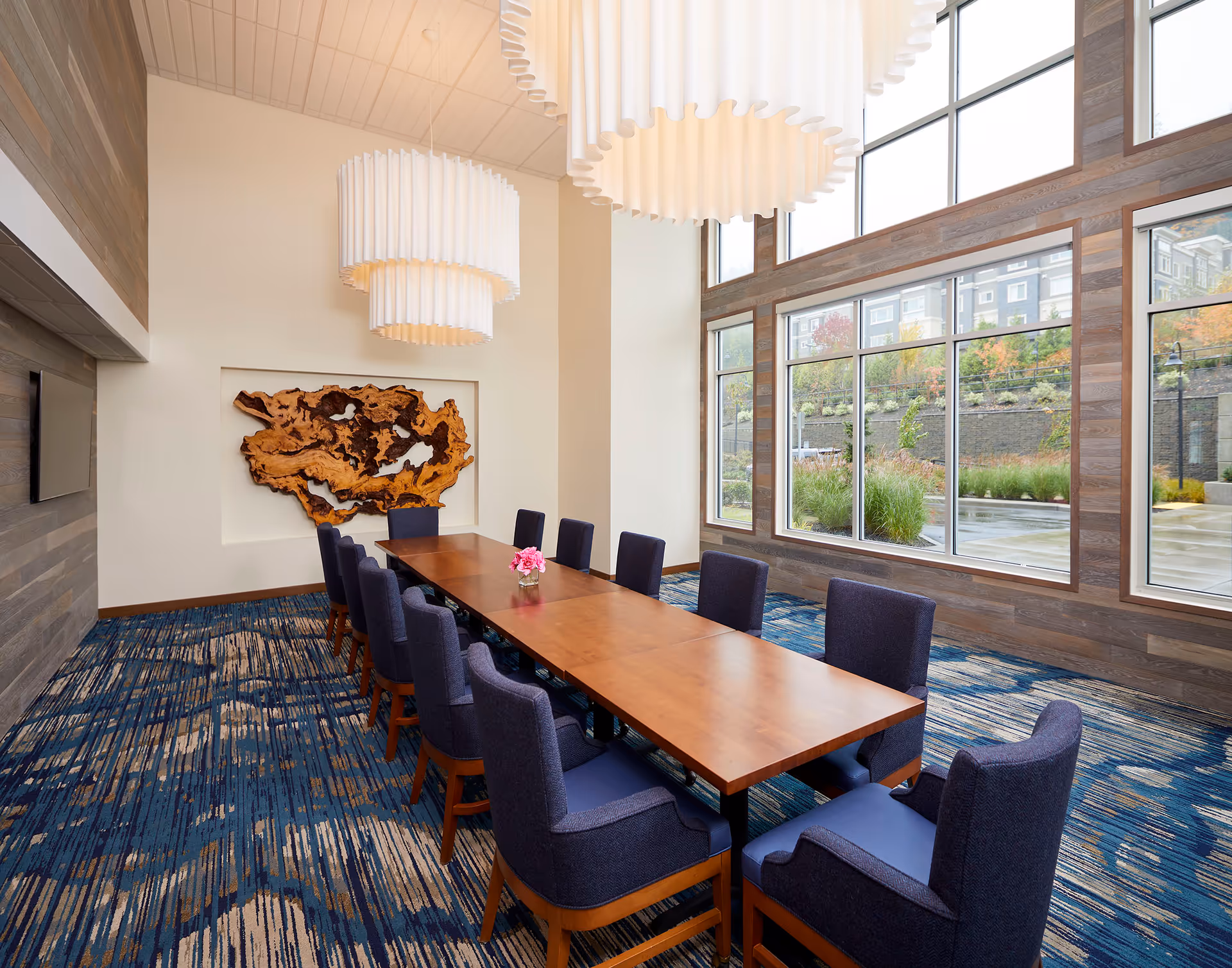 A long wooden table with blue chairs sits in a bright dining room with large windows.