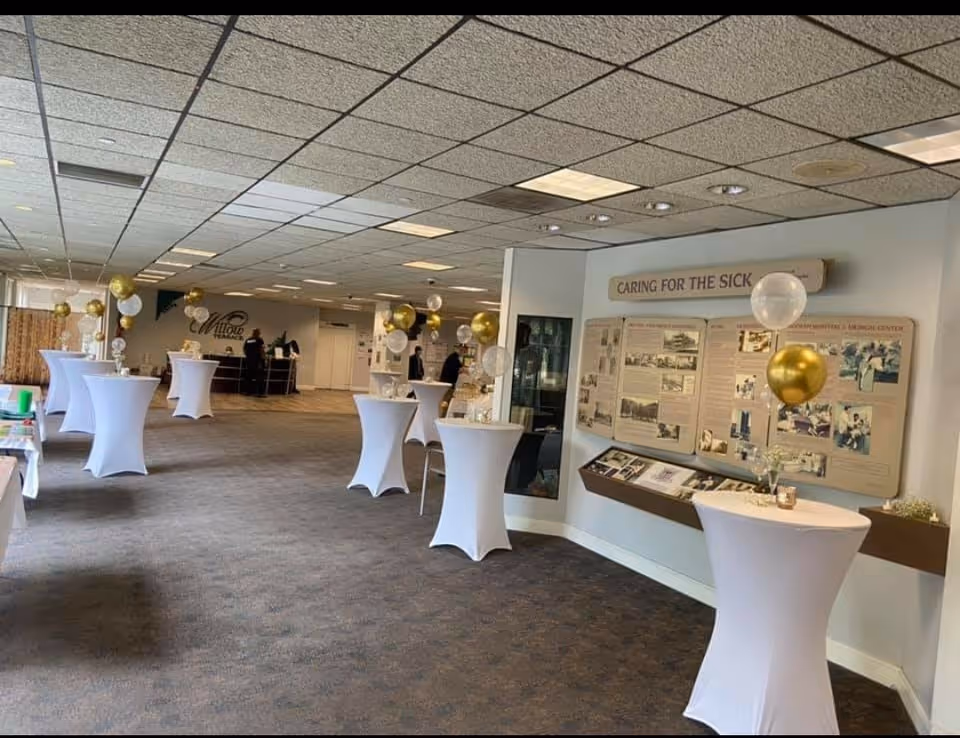 Interior view of a large room set up for an event at Willow Terrace, featuring tall cocktail tables covered with white cloths and decorated with gold and white balloons. The room has a carpeted floor, a drop ceiling with fluorescent lights, and informational displays on the wall titled 'Caring for the Sick.' A few people are visible in the background near the reception area.