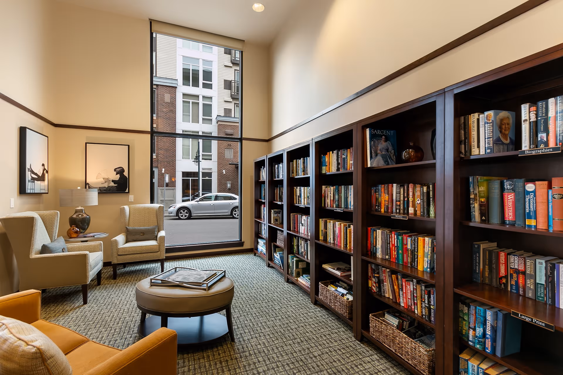 A cozy library or reading room with tall bookshelves filled with books along one wall, two beige armchairs with a small table and lamp between them, a round ottoman in the center, and a large window showing a street and building outside.