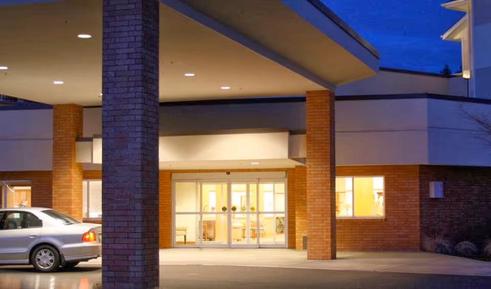 Entrance of Friendsview Retirement Community at dusk with a covered drop-off area supported by brick pillars, illuminated interior visible through glass doors and windows, and a silver car parked nearby.