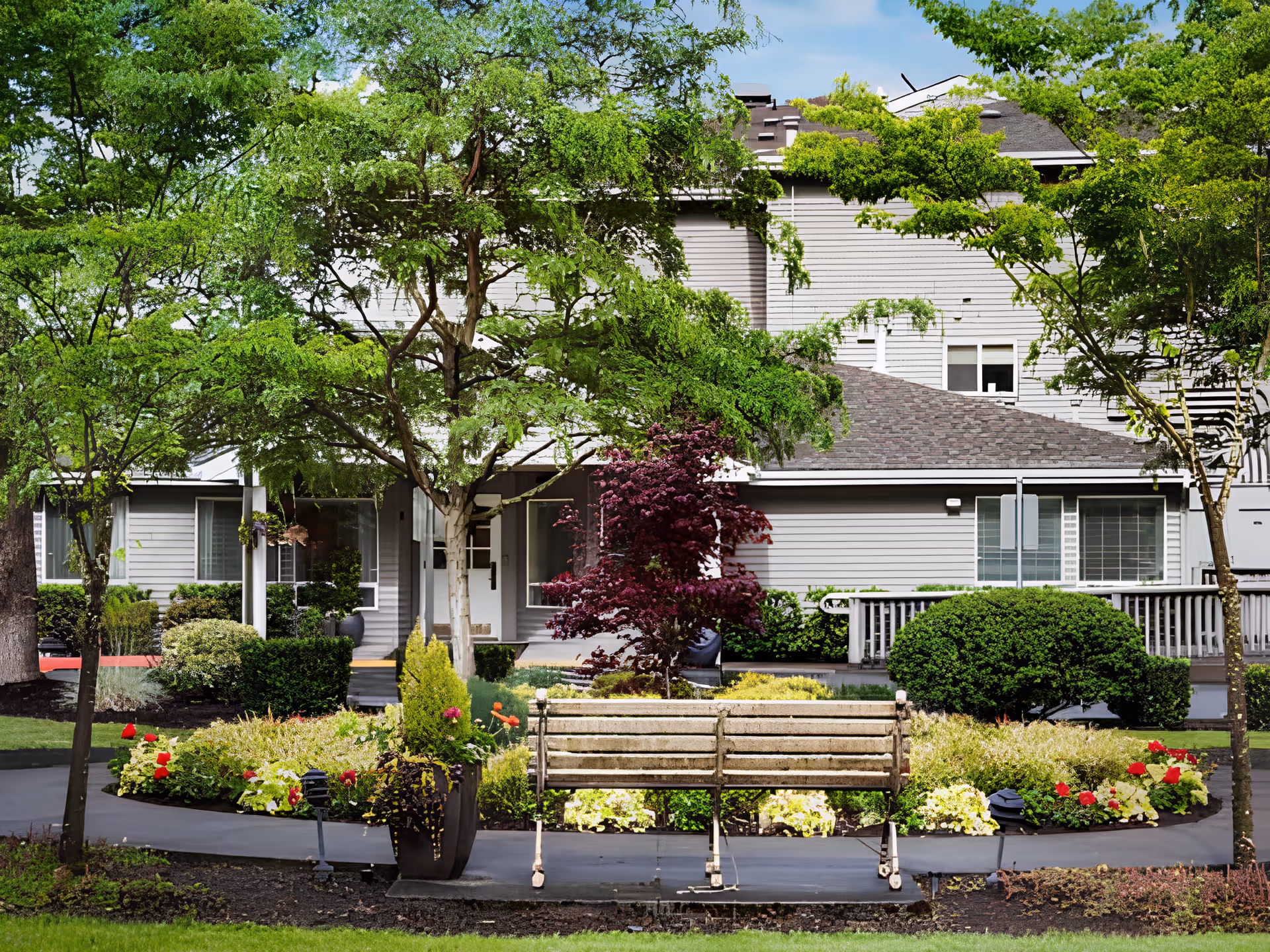 A peaceful outdoor garden area at Cogir of Bothell Memory Care featuring a wooden bench surrounded by colorful flowers, green shrubs, and trees with a gray building in the background.