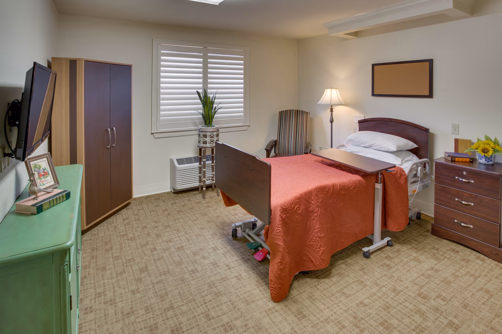 A furnished senior bedroom with a single hospital-style bed covered by a red bedspread, a bedside table and lamp, dresser, TV, and a window with shutters.