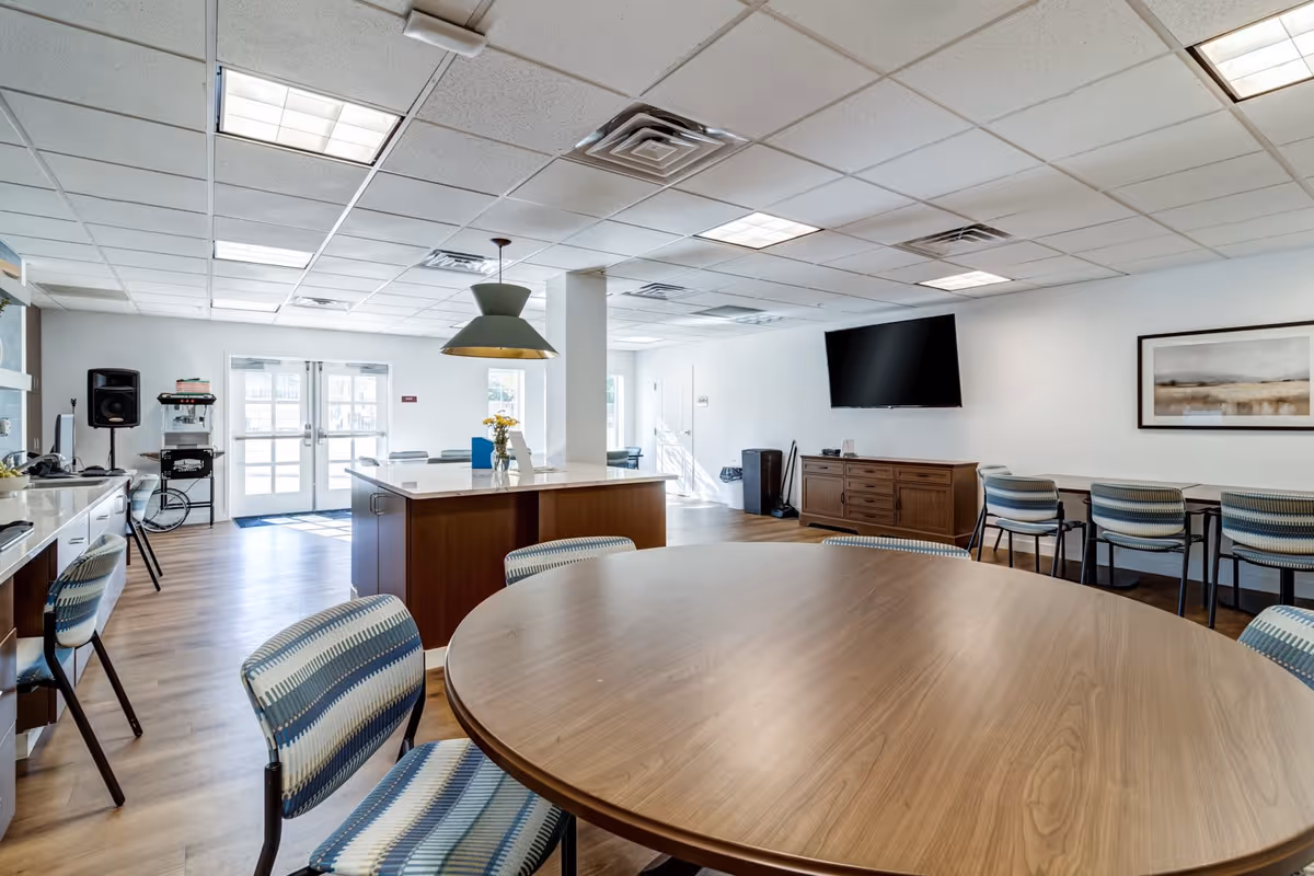 A bright and spacious common area in a senior living facility featuring a large round wooden table with striped chairs around it. In the background, there is a kitchen island with a hanging light fixture above it, a wall-mounted flat-screen TV, a wooden sideboard, and additional seating along the wall. The room has large windows and double glass doors allowing natural light to enter, with light-colored walls and a wood floor.