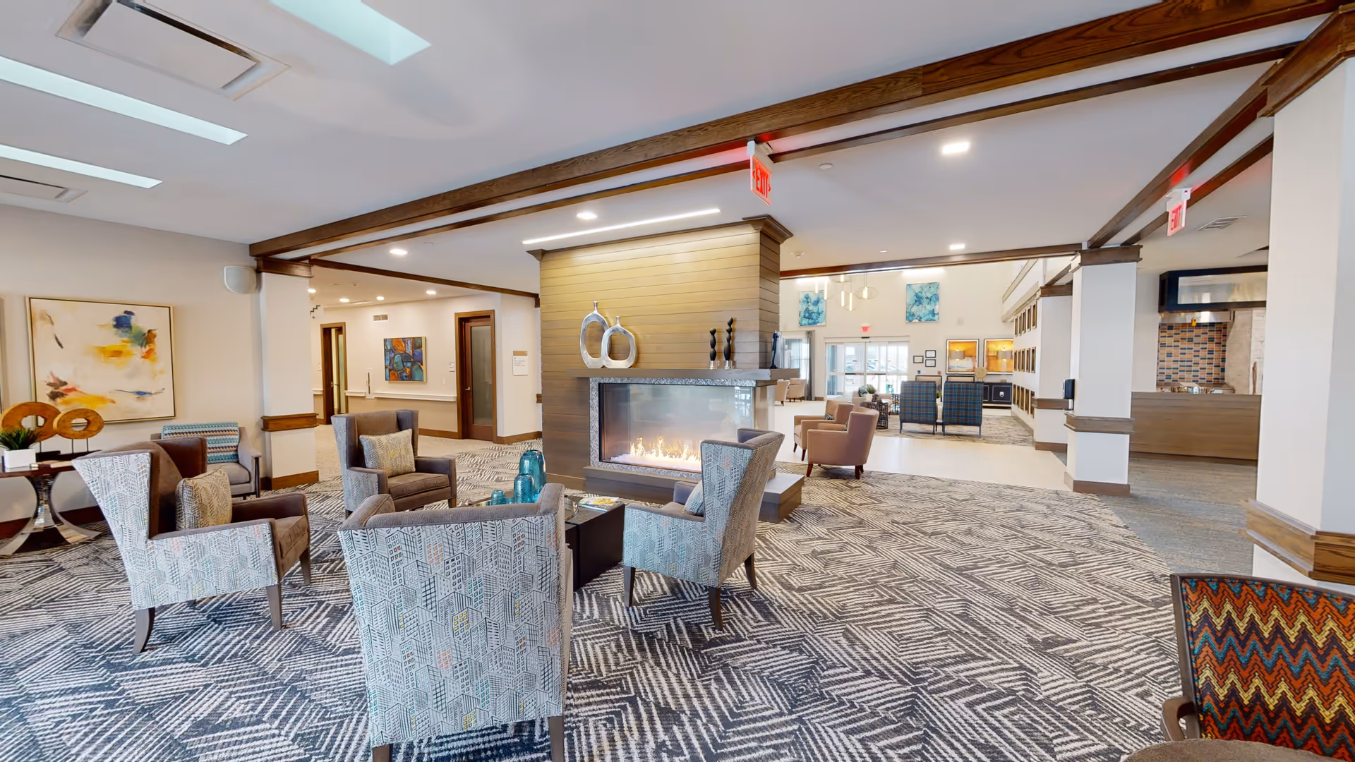 A spacious senior living facility common area with patterned carpet, multiple armchairs arranged around a modern double-sided fireplace, decorative vases on the mantel, and colorful abstract artwork on the walls. The area is well-lit with recessed lighting and skylights, and there is a reception or dining area visible in the background.