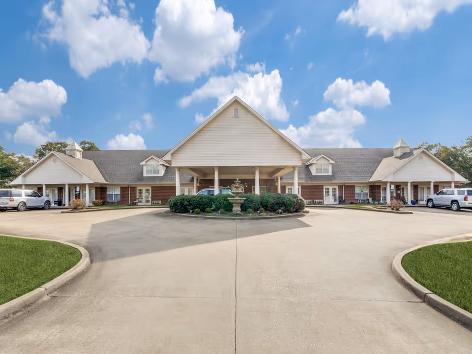 Front exterior view of Cypress Place facility showing a large driveway with a circular landscaped area featuring a fountain in the center. The building has a brick lower facade with white siding on the upper part and a covered entrance supported by columns. Several vehicles are parked on either side of the driveway under smaller covered areas. The sky is blue with scattered clouds.