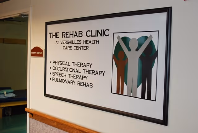 A framed sign on a wall inside a healthcare facility that reads 'THE REHAB CLINIC AT VERSAILLES HEALTH CARE CENTER' listing services including Physical Therapy, Occupational Therapy, Speech Therapy, and Pulmonary Rehab. To the right of the text is a graphic of three stylized human figures with arms raised in front of a heart shape. A small sign on the adjacent wall reads 'THERAPY SERVICES'.