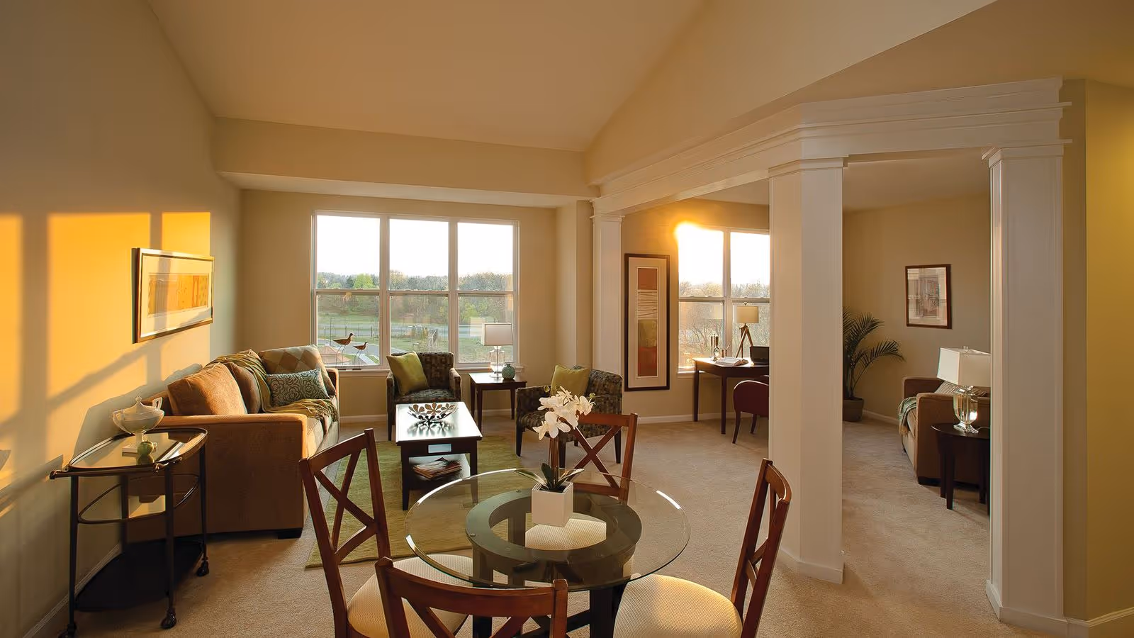 A bright and spacious living area in a senior living community featuring a glass-top dining table with four wooden chairs in the foreground. The room has beige walls and carpet, a comfortable seating area with sofas and armchairs near large windows letting in natural light, and a small desk with a chair in the background. Decorative elements include framed artwork, table lamps, and a potted plant.
