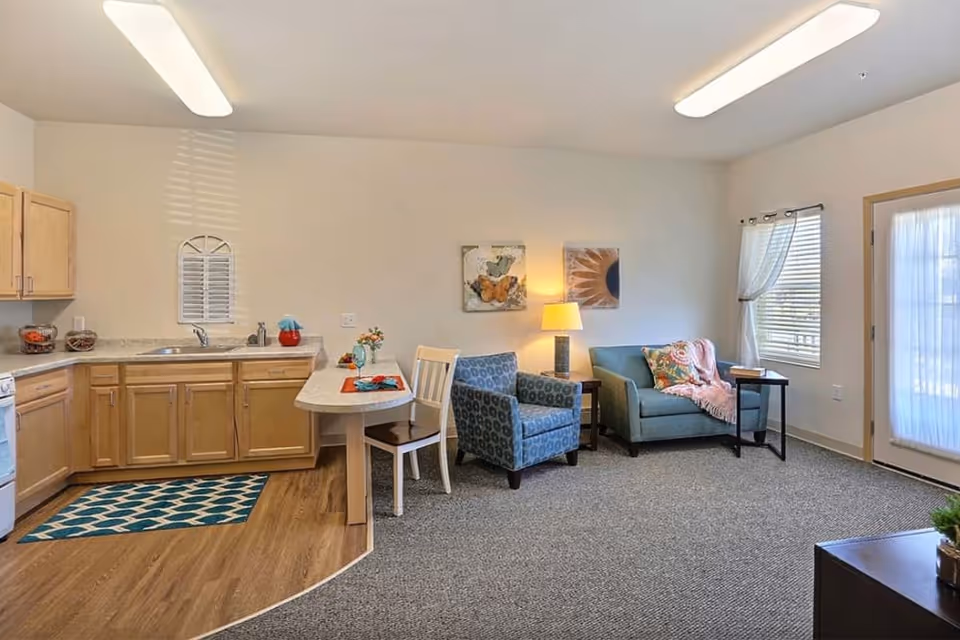 Bright studio-style living area with a small kitchenette, dining counter, upholstered armchair and loveseat beside a lamp and a door to the outside.