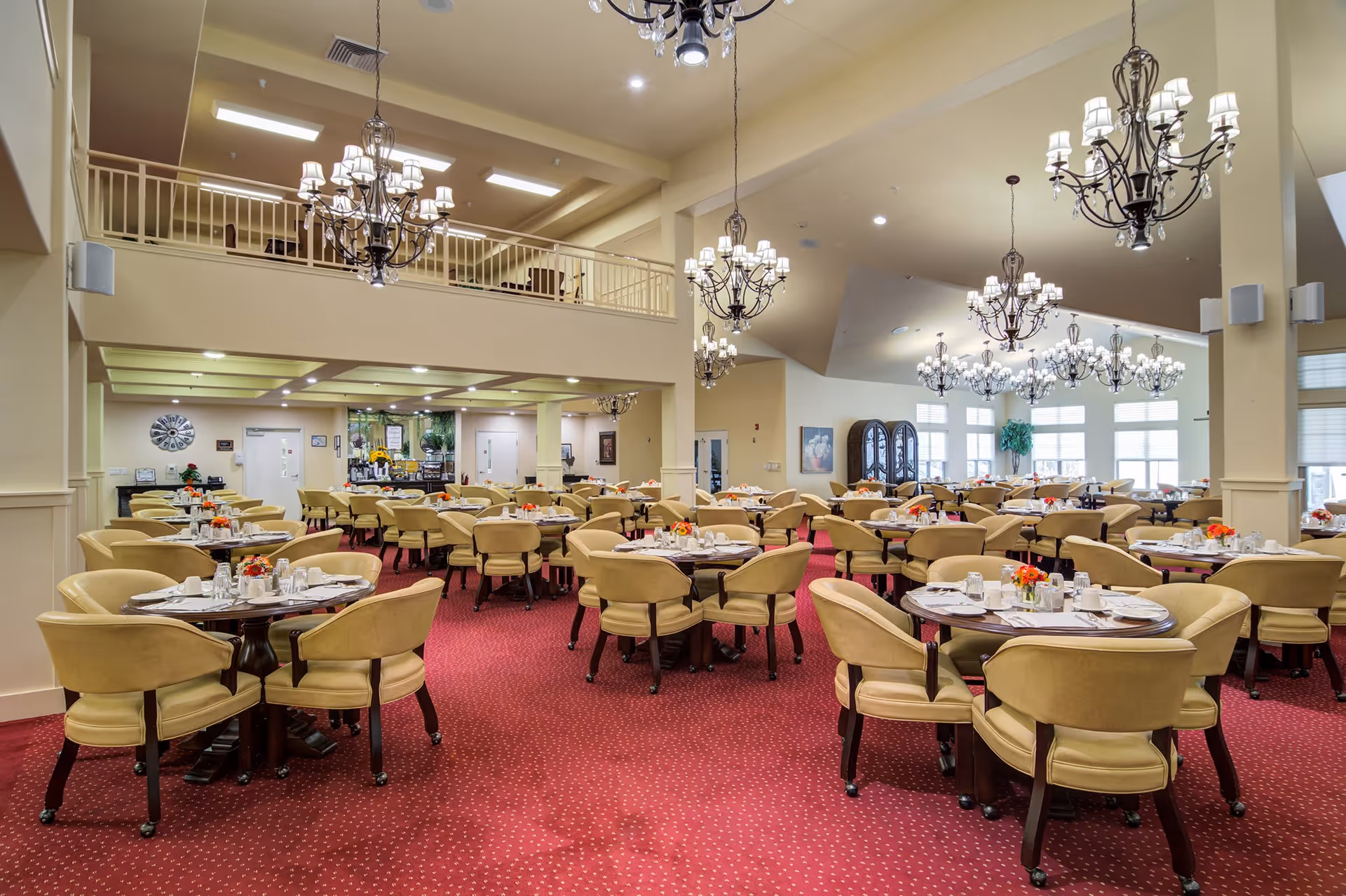 A spacious dining room with multiple round tables, each surrounded by beige upholstered chairs. The tables are set with white napkins, glassware, and small floral centerpieces. The room features red carpet flooring, cream-colored walls, large windows allowing natural light, and several ornate chandeliers hanging from the ceiling. There is a balcony area above and some decorative elements on the walls.