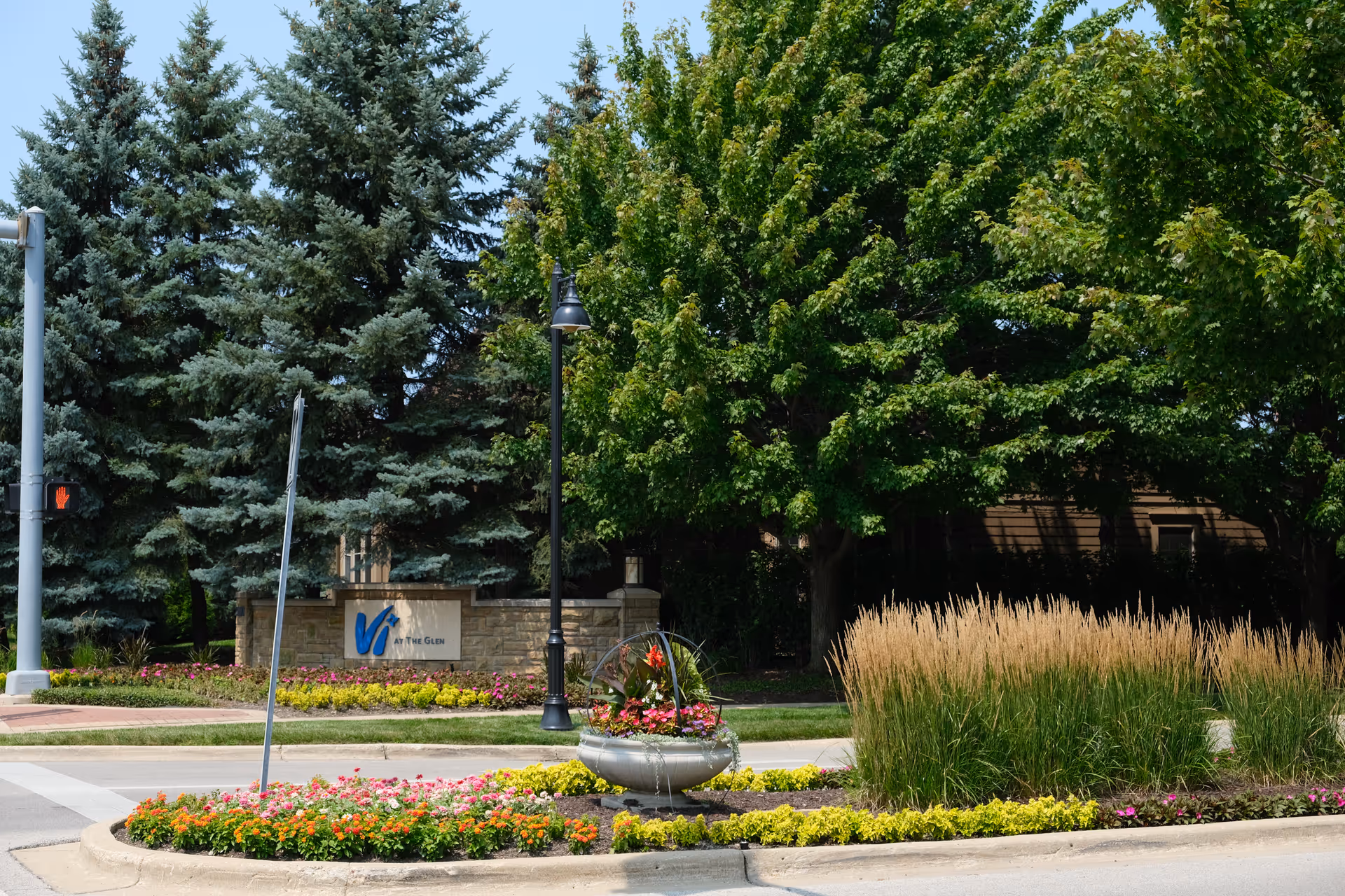 Landscaped entrance with trees, flowerbeds, a lamp post, and a stone sign reading "Vi at The Glen".