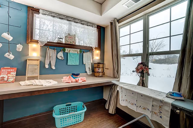 A bright laundry room with a countertop holding folded clothes and a small washboard. Above the counter, a window with lace curtains and a decorative sign that says 'LAUNDRY' with small clothes hanging on a string. Below the counter is a turquoise laundry basket. To the right, an ironing board with a floral cloth and an iron sits in front of a large window with beige curtains, showing a snowy outdoor scene.