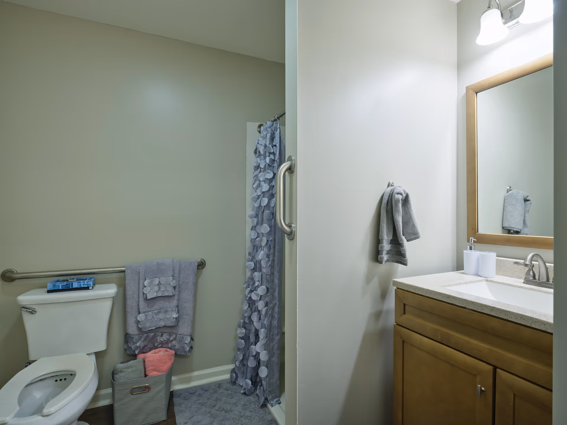 A bathroom with a toilet, a shower with a gray patterned curtain, a towel rack with gray towels, a gray bath mat, a basket with towels, and a wooden vanity with a sink, mirror, and soap dispenser.