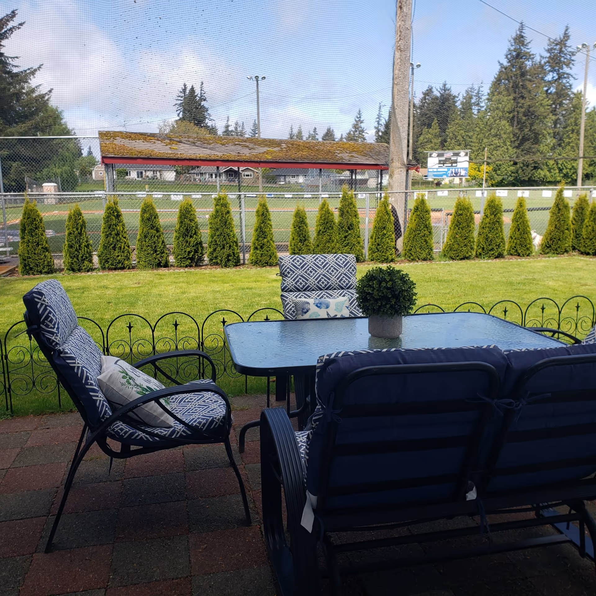 Outdoor patio area with a glass table and cushioned chairs on a tiled floor. Beyond the patio is a green lawn bordered by a small decorative fence, with a row of tall shrubs and a chain-link fence behind them. In the background, there is a sports field with a covered dugout and trees under a partly cloudy sky.