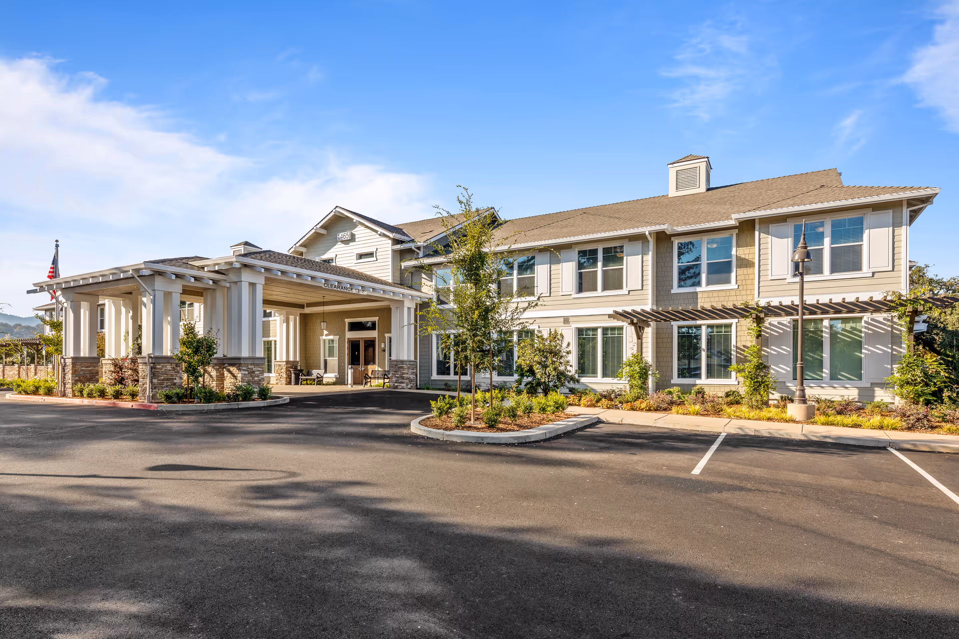 Exterior view of a two-story senior living facility building with a covered entrance, landscaped greenery, and a clear blue sky.