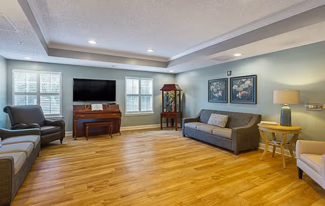 A spacious living room with wooden flooring, light blue walls, and recessed ceiling lights. The room features a gray sofa with a decorative pillow, two armchairs, a wooden piano with a bench, a flat-screen TV mounted on the wall between two windows with white shutters, a small wooden table with a lamp, and two framed floral paintings on the wall.
