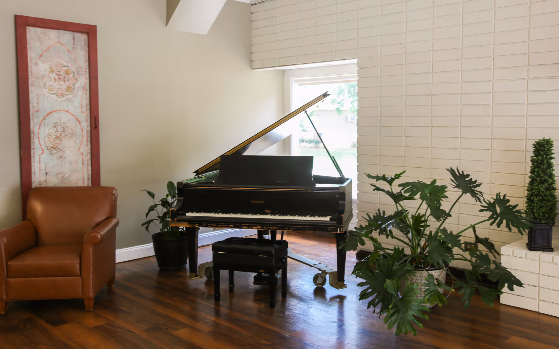 A bright communal living room area featuring a black grand piano, a leather armchair, and several potted plants against a white brick wall.