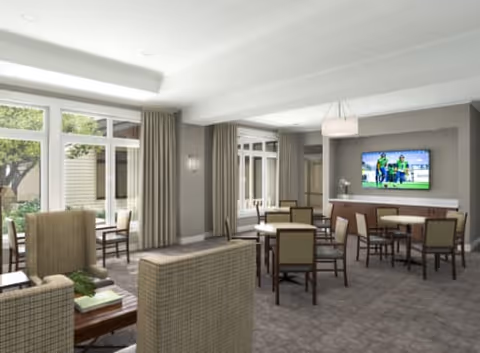 A spacious senior living common area with multiple tables and chairs arranged for socializing or dining. The room features large windows with beige curtains allowing natural light to enter. There is a flat-screen TV mounted on the wall displaying a sports game. The decor is neutral with beige and brown tones, and the carpeted floor adds warmth to the space.