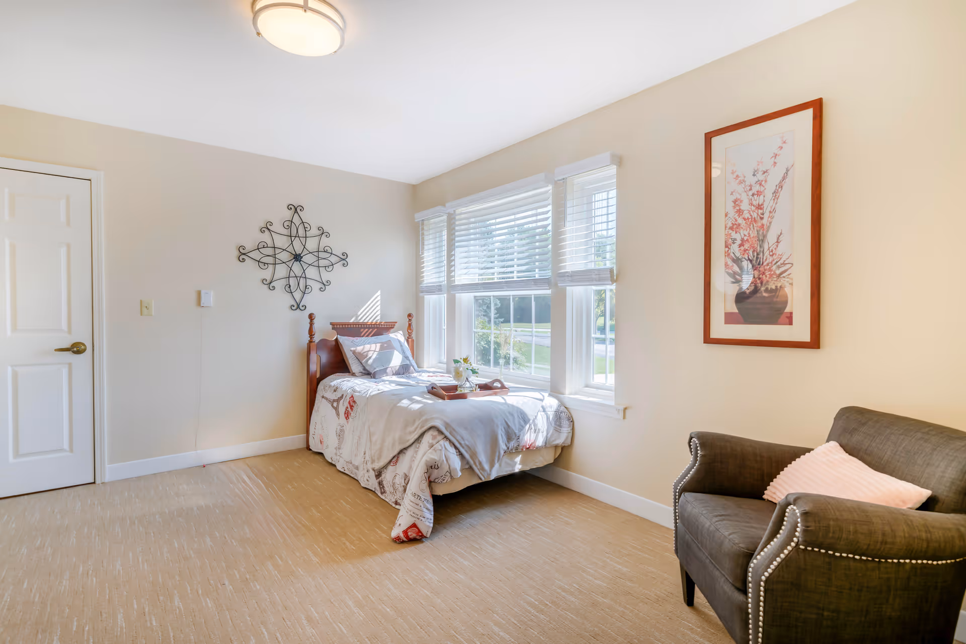 A bright bedroom with a single bed placed near a window with white blinds. The bed has a light-colored comforter and pillows, and a tray with a small plant is on the bed. There is a dark upholstered armchair with a pink pillow on the right side and a framed floral artwork hanging above it. The walls are light beige, and there is a decorative metal wall hanging above the bed. A closed white door is visible on the left side of the room.