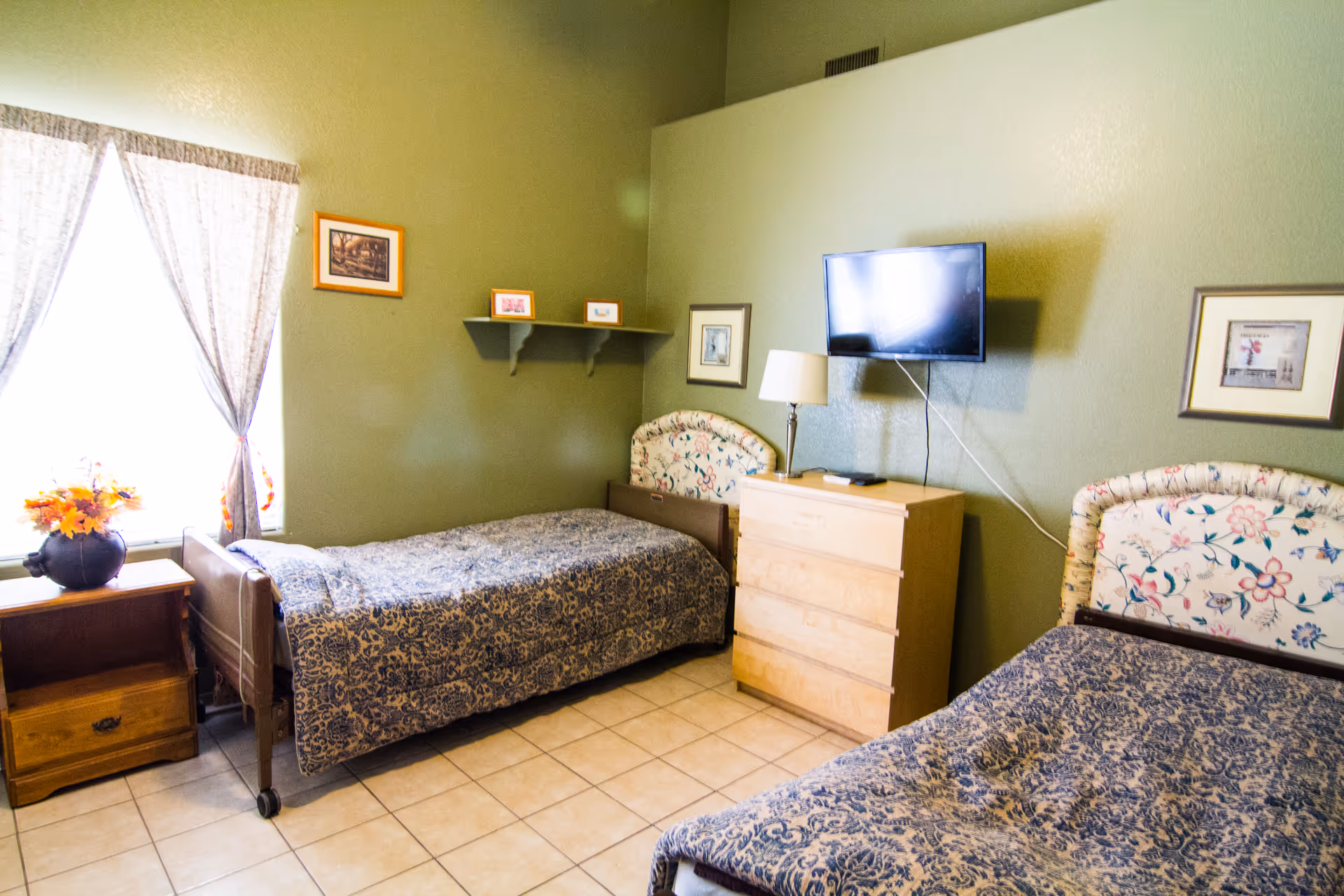 A bedroom with two single beds featuring floral-patterned headboards and blue patterned bedspreads. There is a wooden nightstand with a vase of flowers next to one bed, a wooden dresser with a lamp and a wall-mounted TV above it, and framed pictures on the green walls. A window with sheer curtains allows natural light into the room.