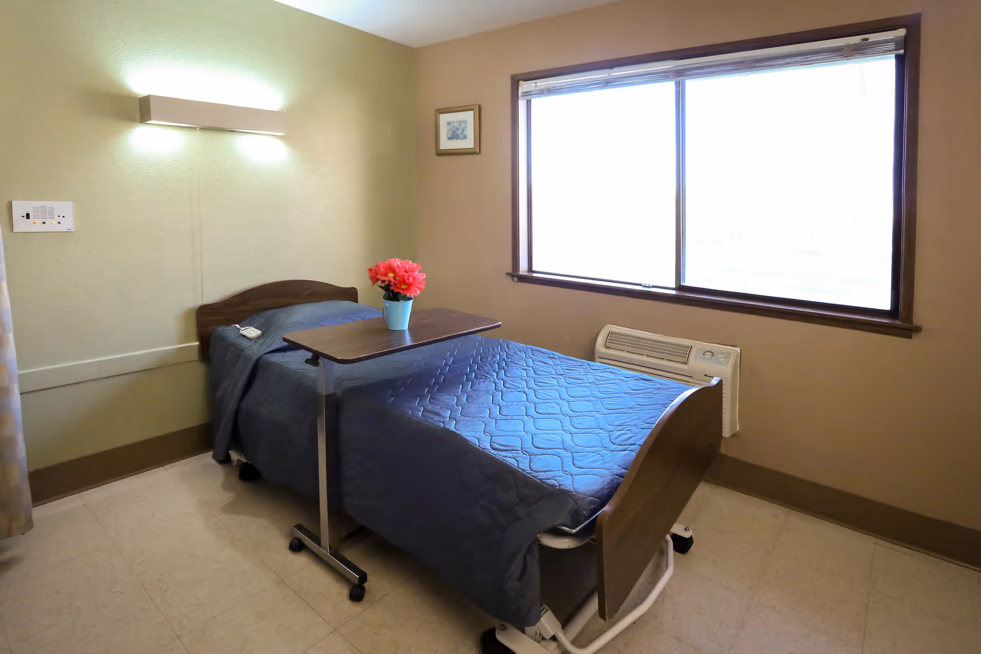 A small bedroom in a senior living facility with a single hospital-style bed covered with a blue quilt. A small overbed table with a vase of red flowers is positioned over the bed. There is a large window with blinds on the right wall and a wall-mounted light fixture above the bed. The room has beige walls and a tiled floor.