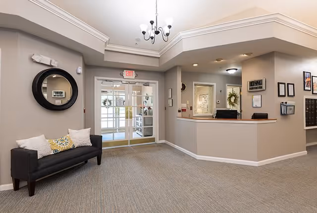 Interior view of a senior living facility lobby area with a reception desk on the right, a black cushioned bench with decorative pillows on the left, a round mirror above the bench, and glass double doors leading outside. The walls are painted light gray and the ceiling has a chandelier light fixture.