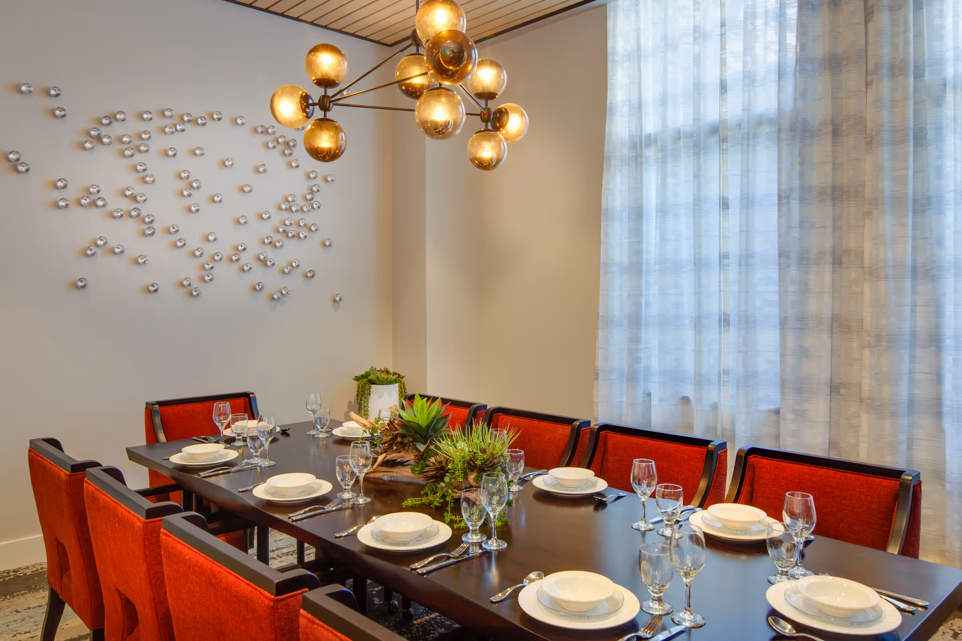 A dining room with a long dark wooden table set for eight people with white plates, bowls, silverware, and glassware. The chairs have red upholstery. There is a modern chandelier with round bulbs hanging above the table. A decorative wall installation with small metallic spheres is on the left wall, and sheer curtains cover a large window on the right.
