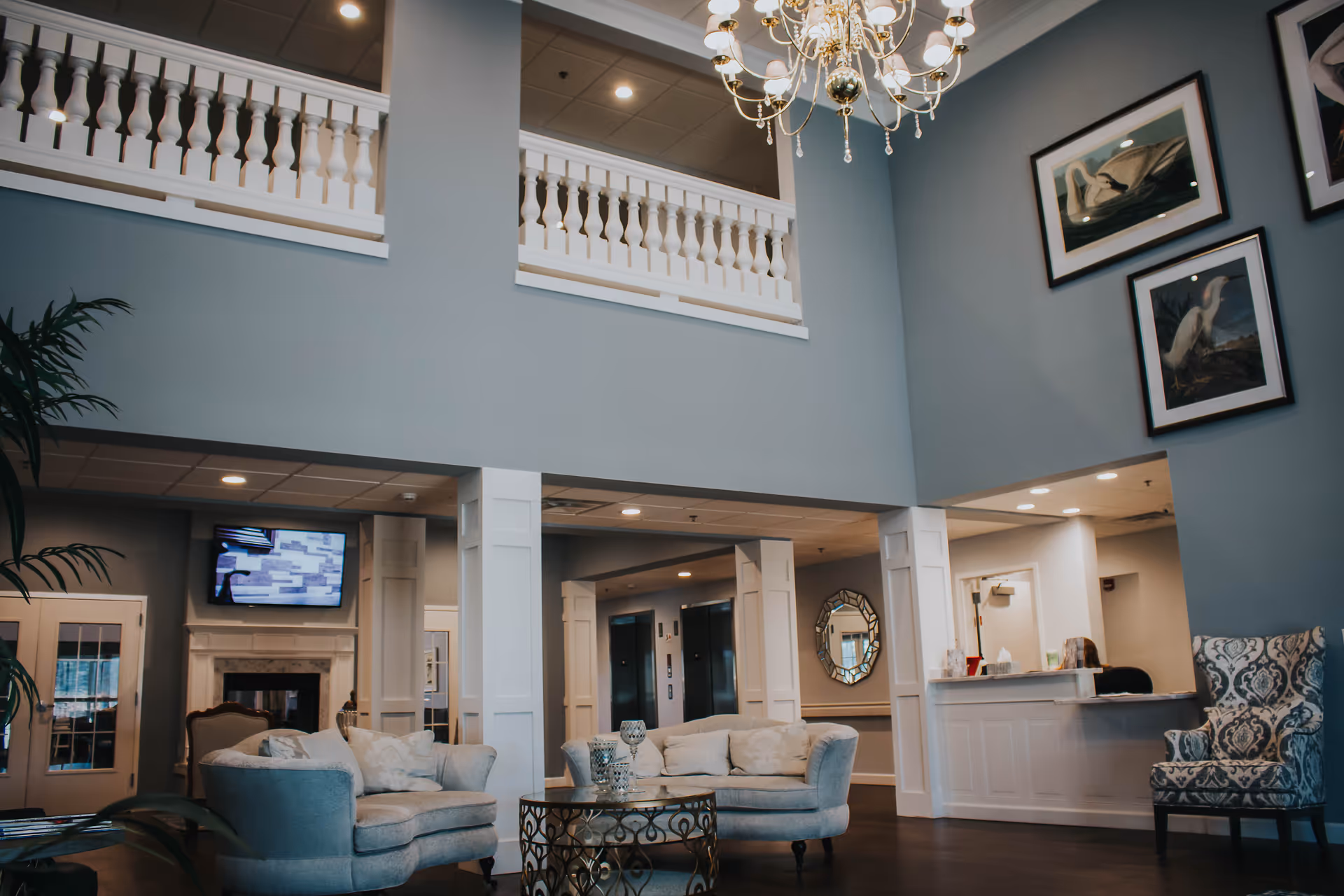 A spacious and elegant senior living facility common area with high ceilings, a chandelier, comfortable upholstered chairs and sofas, a decorative round coffee table, framed bird artwork on the walls, a fireplace with a TV mounted above it, and a reception desk in the background.
