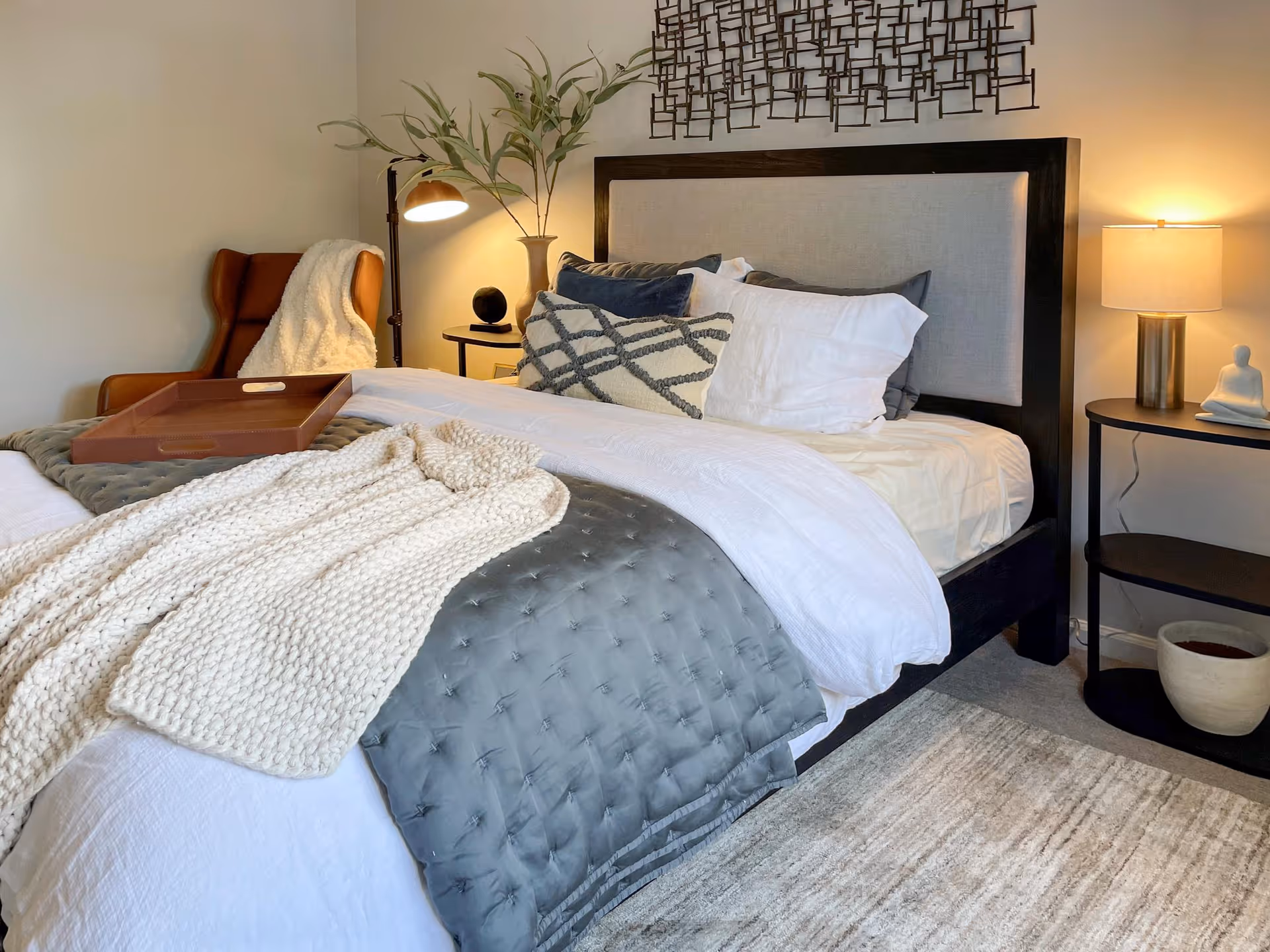 Sunlit bedroom with a made bed, textured throw and pillows, a tray, armchair, and bedside tables with lamps.