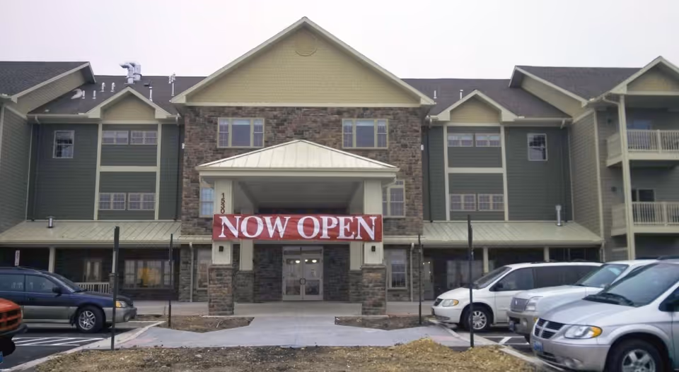 Front exterior view of a multi-story building with a stone and green siding facade. A large red banner with white text reading 'NOW OPEN' hangs above the main entrance. Several parked cars are visible in the parking lot in front of the building.