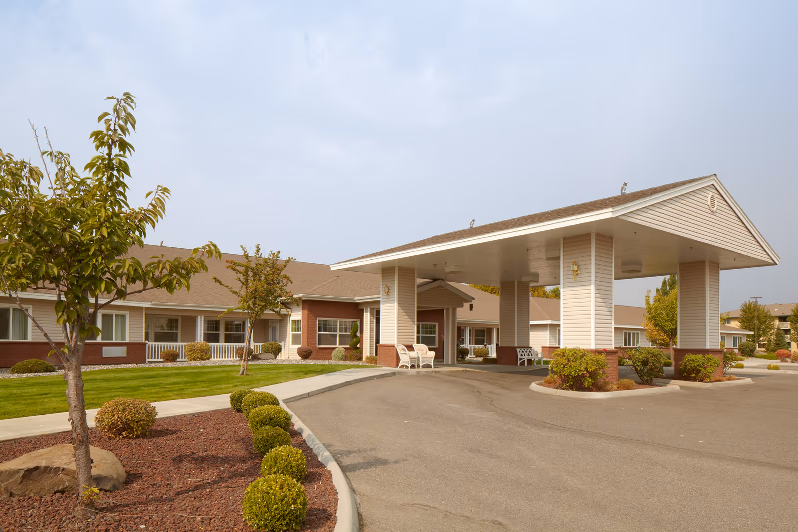 Porte-cochère entrance of a single-story senior living facility with landscaped grounds, shrubs, and a circular driveway.