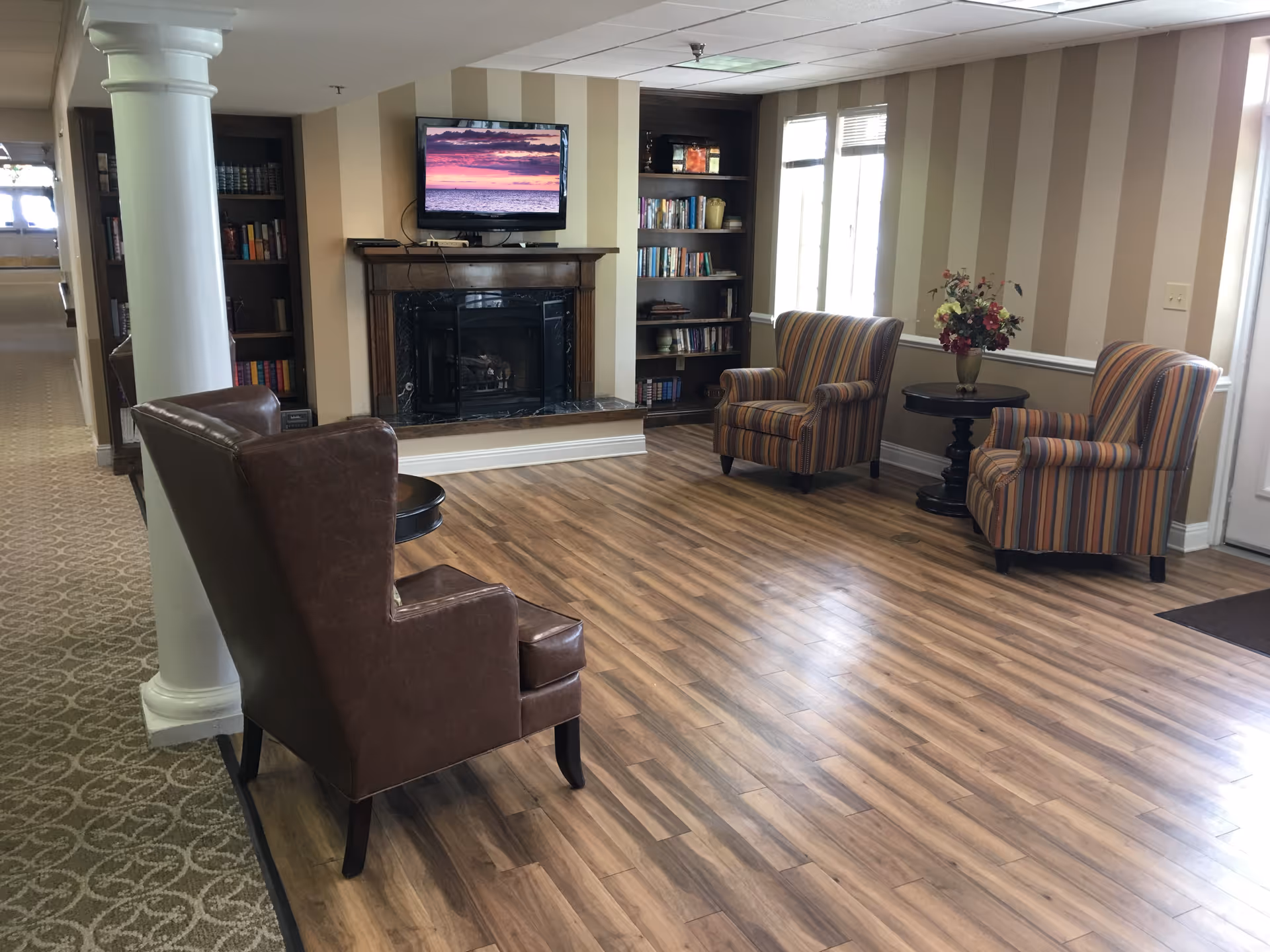 A cozy living room area with a wooden floor, featuring a brown leather armchair and two striped upholstered armchairs around a small round table with a flower vase. There is a fireplace with a TV mounted above it displaying a sunset over the ocean. Bookshelves flank the fireplace, and large windows allow natural light to fill the room.