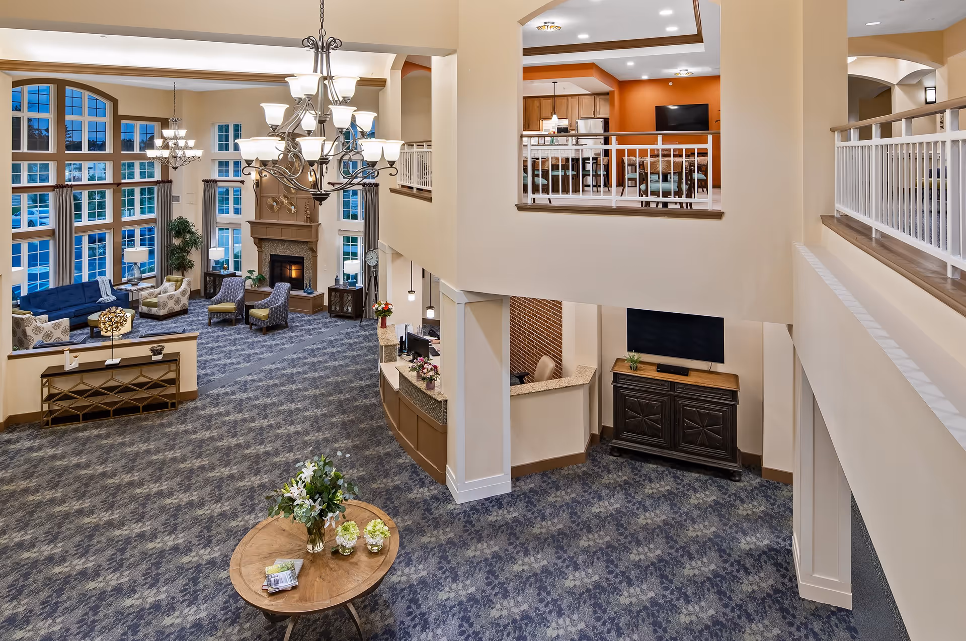 Spacious senior living facility common area with high ceilings and large windows. The room features a seating area with sofas and armchairs arranged around a fireplace, a chandelier hanging from the ceiling, a reception desk, and an upper-level dining area with tables and chairs. The decor includes neutral walls, patterned carpet, and floral arrangements on a round wooden table.