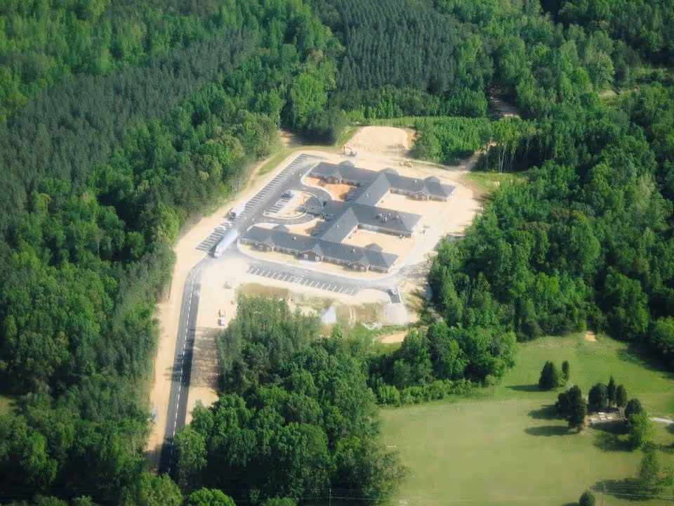 Aerial view of Caswell House, a large building complex surrounded by dense green forest and open grassy areas, with a paved road leading to the parking lot.